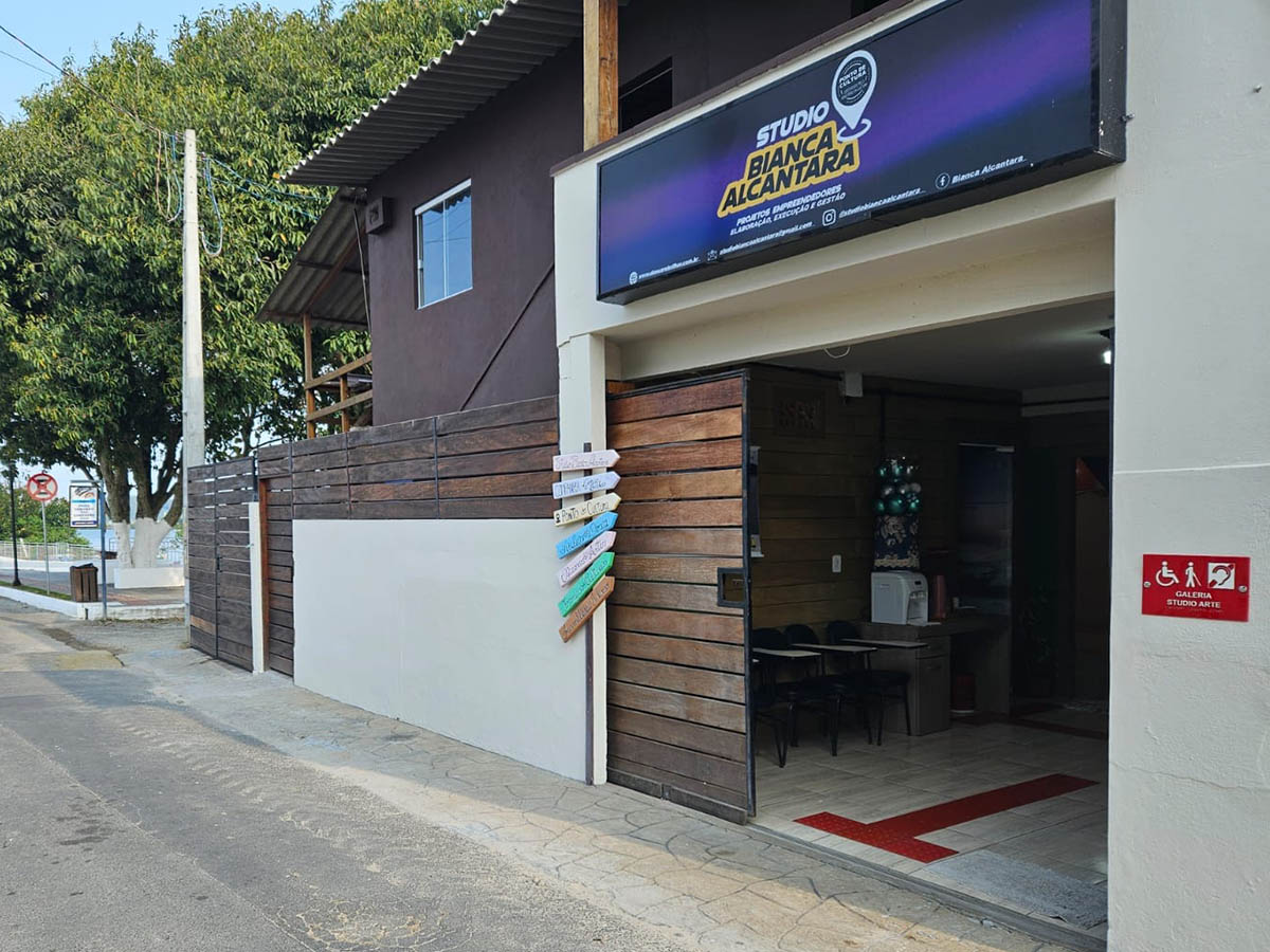 À frente, há uma entrada com um portão de madeira marrom e preto. Acima do portão há uma placa roxa com o logo do Studio Bianca Alcantara: a palavra "studio" em letras maiúsculas, brancas e Bianca Alcantara em letras pretas sobre um fundo amarelo. À direita há um ícone de localização branco sobre um fundo preto, com a inscrição Ponto de Cultura certificado pelo Ministério da Cultura. Abaixo, em letras brancas: projetos empreendedores, elaboração, execução e gestão. Na entrada do studio há um piso podotátil vermelho que vai em direção a algumas cadeiras e uma mesa, um filtro de água, uma sala de atendimento e uma sala multicultural. Do lado de fora, em uma parede à direita do studio, há uma placa em braille, vermelha, com a frase: Galeria Studio Arte. À esquerda da entrada há uma placa de madeira com flechas rosa, azul, amarela, verde e marrom apontando para a entrada. À esquerda do Studio há uma casa com portão de madeira marrom e uma praça com uma grande árvore. FIM DA AUDIODESCRIÇÃO.